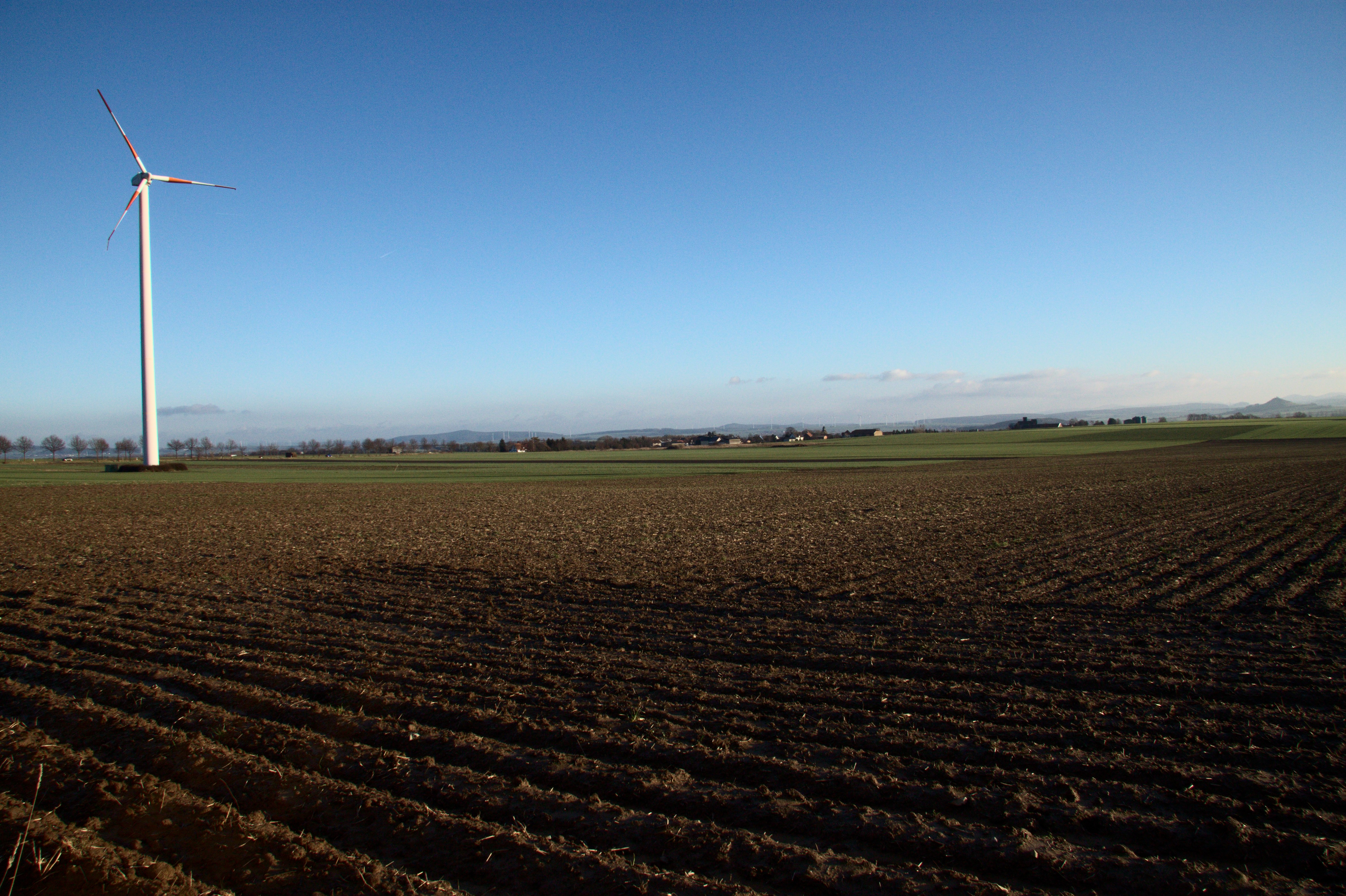 Blick über ein geackertes Feld in die Ebene der Warburger Börde. Links eine Windkraftanlage, am Horizont das Dorf Eissen und rechts in der Ferne der Desenberg.