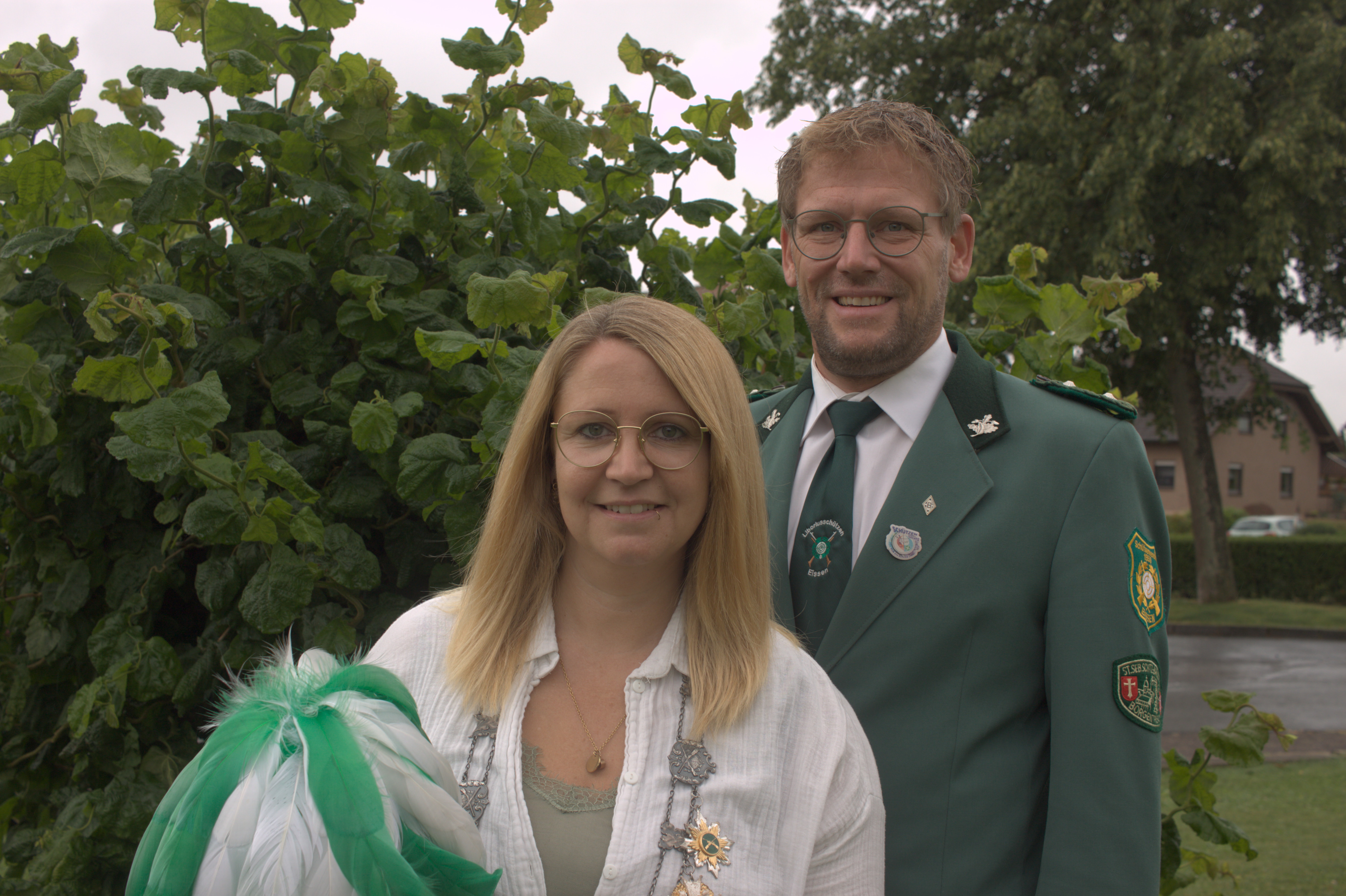 Eine Frau und ein Mann stehen vor einem grünen Busch in einem Garten. Die Frau hat eine Königskette um den Hals und hält einen Hut in der Hand. Der Mann trägt eine grüne Uniform.